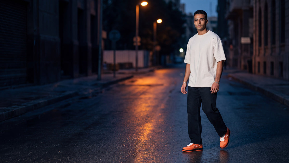 A man walking across an empty wet city street at blue hour in orange Nike Dunks, cream oversized tee and black trousers, streetlights reflected on the asphalt
