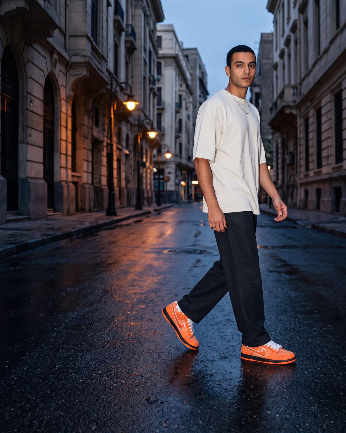 A man walking across an empty wet city street at blue hour in orange Nike Dunks, cream oversized tee and black trousers, streetlights reflected on the asphalt
